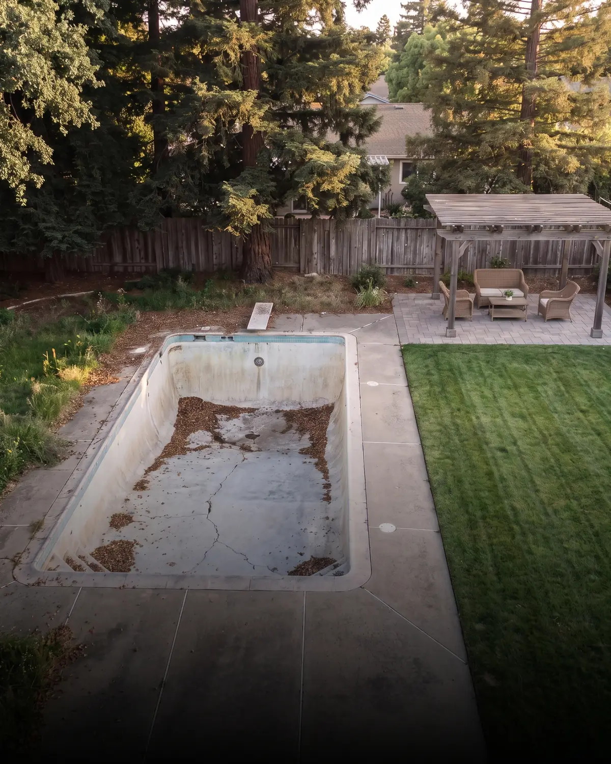 An aerial view of one Sacramento backyard shown mid-transformation — an old empty concrete pool with cracked plaster and a weed-cracked deck, and the same yard already reclaimed as mown lawn, drought planting and a paved patio under a wooden pergola.